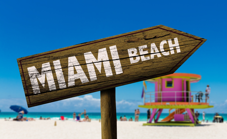 Wooden sign with MIAMI BEACH printed on it, with a beach, ocean, and lifeguard tower