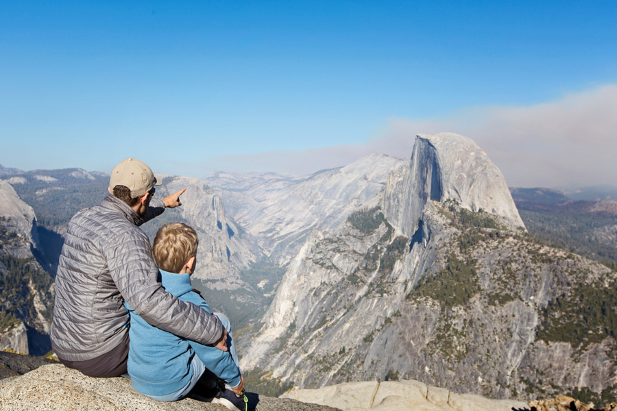 back view of father and son sitting together at glacier point and enjoying view of half dome and yosemite valley in yosemite national park, california in december 